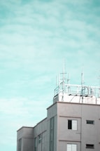 A modern building features several antennas and communication equipment on its rooftop against a cloudy sky. The structure is composed of rectangular windows with shutters, and the overall design is clean with a minimalist aesthetic.