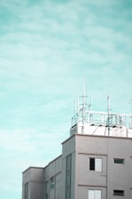 A modern building features several antennas and communication equipment on its rooftop against a cloudy sky. The structure is composed of rectangular windows with shutters, and the overall design is clean with a minimalist aesthetic.