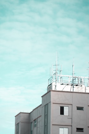 A modern building features several antennas and communication equipment on its rooftop against a cloudy sky. The structure is composed of rectangular windows with shutters, and the overall design is clean with a minimalist aesthetic.