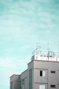 A modern building features several antennas and communication equipment on its rooftop against a cloudy sky. The structure is composed of rectangular windows with shutters, and the overall design is clean with a minimalist aesthetic.