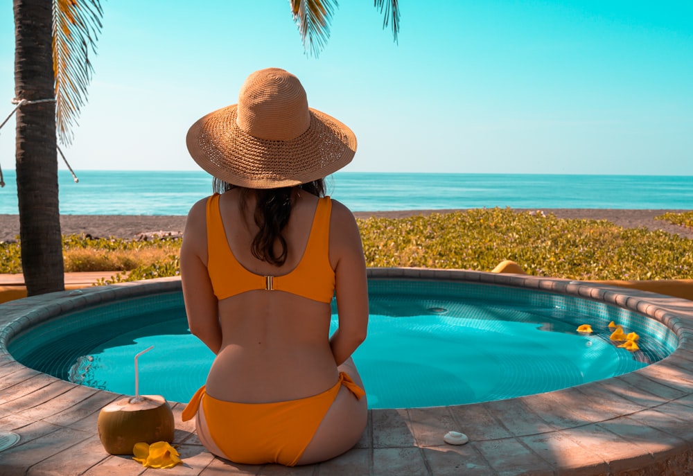 Frau mit braunem Sonnenhut und blauem Bikinihöschen sitzt tagsüber auf dem Swimmingpool