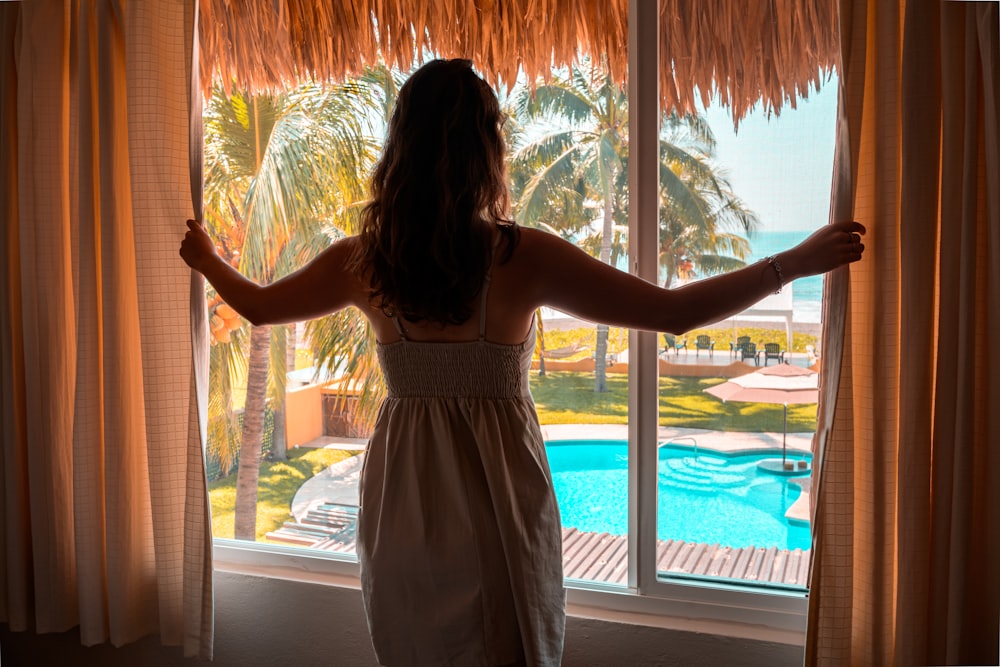 woman in white spaghetti strap dress standing near window during daytime