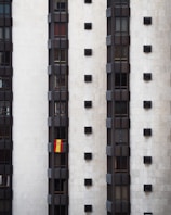 A tall, modern building facade with multiple vertical sections of dark windows is shown. A Spanish flag hangs prominently from one of the windows, adding a splash of color to the otherwise monochromatic scene.