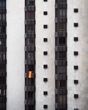 A tall, modern building facade with multiple vertical sections of dark windows is shown. A Spanish flag hangs prominently from one of the windows, adding a splash of color to the otherwise monochromatic scene.
