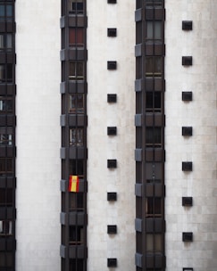 A tall, modern building facade with multiple vertical sections of dark windows is shown. A Spanish flag hangs prominently from one of the windows, adding a splash of color to the otherwise monochromatic scene.