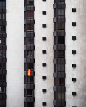 A tall, modern building facade with multiple vertical sections of dark windows is shown. A Spanish flag hangs prominently from one of the windows, adding a splash of color to the otherwise monochromatic scene.