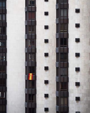 A tall, modern building facade with multiple vertical sections of dark windows is shown. A Spanish flag hangs prominently from one of the windows, adding a splash of color to the otherwise monochromatic scene.