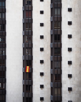 A tall, modern building facade with multiple vertical sections of dark windows is shown. A Spanish flag hangs prominently from one of the windows, adding a splash of color to the otherwise monochromatic scene.