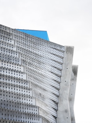 A futuristic building facade with a series of perforated metallic panels arranged in a layered, geometric fashion. The panels have a matte gray finish with rows of uniformly spaced small circular holes. A section of bright blue is visible at the top, adding a striking contrast to the otherwise monochromatic color scheme.