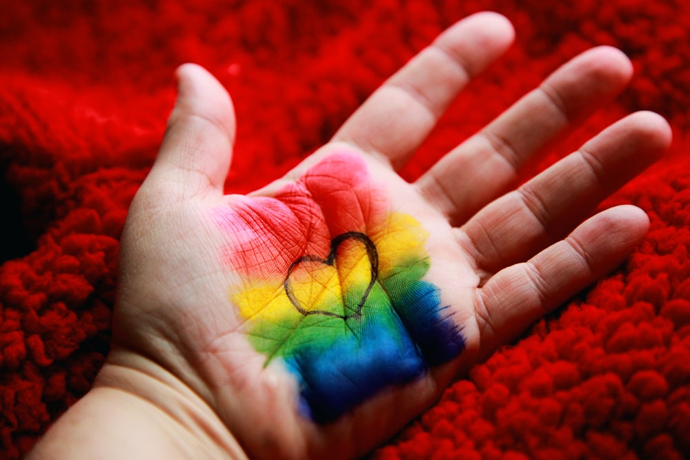 An open palm with rainbow colors drawn on it with markers. A black heart outline is drawn over the colors.