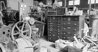 An industrial workshop environment filled with a variety of machinery and tools. In the center, there is a large metal cabinet with multiple drawers, surrounded by old machines and mechanical components. Various pipes, wheels, and cables are scattered around, while sunlight filters in through the windows, casting shadows across the scene.