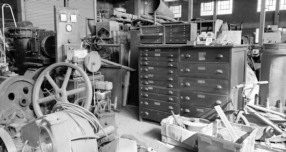 An industrial workshop environment filled with a variety of machinery and tools. In the center, there is a large metal cabinet with multiple drawers, surrounded by old machines and mechanical components. Various pipes, wheels, and cables are scattered around, while sunlight filters in through the windows, casting shadows across the scene.