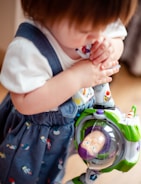 A young child holding a toy Buzz Lightyear figure. The child is wearing a denim-esque jumper with colorful patterns and a white T-shirt. The toy is prominently featured with its green, white, and purple colors.