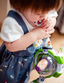 A young child holding a toy Buzz Lightyear figure. The child is wearing a denim-esque jumper with colorful patterns and a white T-shirt. The toy is prominently featured with its green, white, and purple colors.