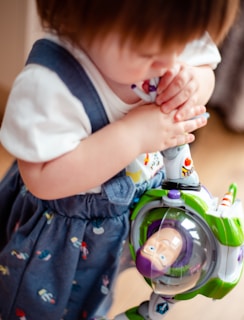 A young child holding a toy Buzz Lightyear figure. The child is wearing a denim-esque jumper with colorful patterns and a white T-shirt. The toy is prominently featured with its green, white, and purple colors.