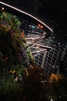 A vibrant vertical garden adorned with lush greenery and colorful flowers, contrasted against a large glass and metal structure. Illuminated walkways and spotlights enhance the modern architectural design, creating a striking interplay between nature and urban elements.
