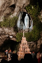 A religious statue of a woman stands in a rocky grotto surrounded by lush greenery. Below, a large candelabrum with numerous lit candles gleams. Several people are gathered, some holding candles, amidst a backdrop of natural stone.