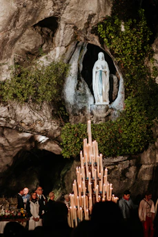A religious statue of a woman stands in a rocky grotto surrounded by lush greenery. Below, a large candelabrum with numerous lit candles gleams. Several people are gathered, some holding candles, amidst a backdrop of natural stone.