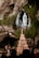A religious statue of a woman stands in a rocky grotto surrounded by lush greenery. Below, a large candelabrum with numerous lit candles gleams. Several people are gathered, some holding candles, amidst a backdrop of natural stone.