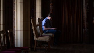 A person is sitting alone on a wooden chair in a dimly lit room with ornate columns and dark curtains. The person appears to be using a phone, and the scene has a quiet, contemplative atmosphere.