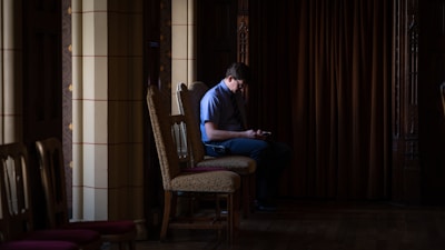A person is sitting alone on a wooden chair in a dimly lit room with ornate columns and dark curtains. The person appears to be using a phone, and the scene has a quiet, contemplative atmosphere.