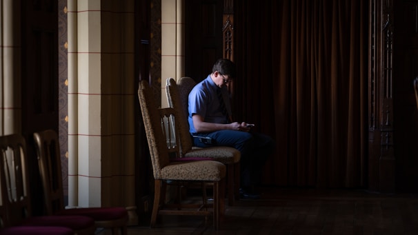 A person is sitting alone on a wooden chair in a dimly lit room with ornate columns and dark curtains. The person appears to be using a phone, and the scene has a quiet, contemplative atmosphere.