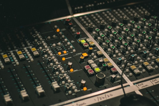 A professional audio mixing console with numerous knobs, sliders, and buttons, featuring an array of colors and lights indicating functionality. Labels with handwritten notes are visible on the board, suggesting customized settings.