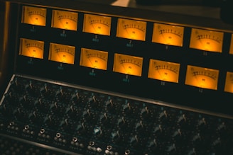 A sound mixing console with multiple VU meters displaying levels and an array of knobs and dials below for audio adjustments. The lighting creates a warm and professional studio atmosphere.