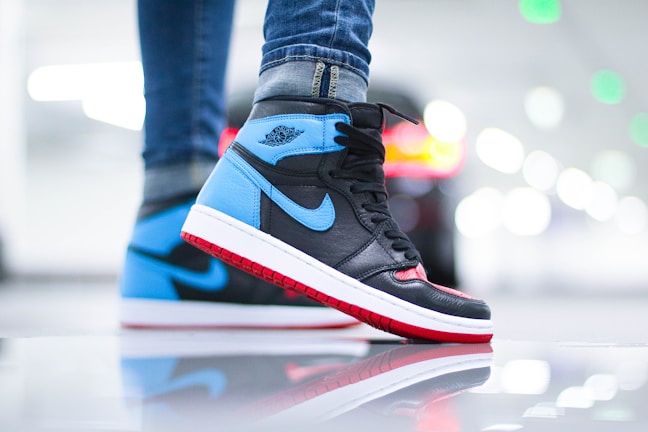 Close-up of a sneaker worn by a person wearing jeans. The sneaker has a distinct design with black, blue, and red colors, featuring a prominent swoosh logo. The background is blurred with circular light bokeh, creating a vibrant and dynamic atmosphere.