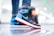 Close-up of a sneaker worn by a person wearing jeans. The sneaker has a distinct design with black, blue, and red colors, featuring a prominent swoosh logo. The background is blurred with circular light bokeh, creating a vibrant and dynamic atmosphere.
