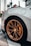 A close-up view of a luxury sports car's rear wheel, showcasing its intricately designed bronze alloy rim and yellow brake caliper. The car has a sleek, metallic body with a focus on the wheel and tire details. A subtle background includes palm trees and buildings, hinting at an urban environment.
