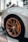 A close-up view of a luxury sports car's rear wheel, showcasing its intricately designed bronze alloy rim and yellow brake caliper. The car has a sleek, metallic body with a focus on the wheel and tire details. A subtle background includes palm trees and buildings, hinting at an urban environment.