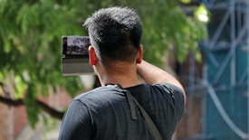 A person with short black hair is holding a smartphone to capture a video or photo. The background features green foliage, suggesting an outdoor setting. The person is wearing a dark shirt and has a shoulder strap hanging on their back.