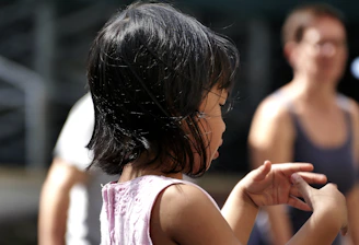 A young child with medium-length black hair is visible in profile, wearing a pink sleeveless top. The child is making a hand gesture, possibly playing or expressing something. In the blurred background, there are two adults, one wearing a white top and the other a dark tank top.