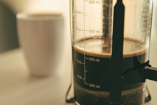 Close-up of a French press coffee maker filled with brewed coffee, with measurement markings visible on the glass. In the blurred background, there is a white coffee cup.