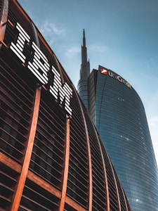 Two prominent skyscrapers with one building featuring a wooden panel design and the IBM logo, while the other shows the UniCredit logo on a sleek glass facade. The sky is clear with a few clouds, creating a modern and corporate atmosphere.