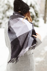 A person is wearing a stylish gray coat and a black winter hat, standing outdoors in a snowy environment. A large, checkered scarf in shades of black, gray, and white is draped around their shoulders.