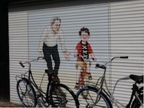 A mural painted on a garage door depicts an older man and a child, presumably a father and son. The child is riding a bicycle with noticeable concentration while the older man is bent over with outstretched arms, suggesting support or guidance. Two real bicycles are parked in front of the mural, blending with the artwork.