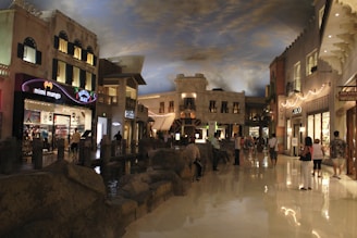 A vibrant indoor shopping area resembling an outdoor street, with multiple storefronts such as 'mimi mango' and 'ALDO'. The floor is reflective and polished, while the ceiling is painted to mimic a cloudy sky. Shoppers are casually walking around, and the area is illuminated by warm, ambient lighting that enhances the architectural features.