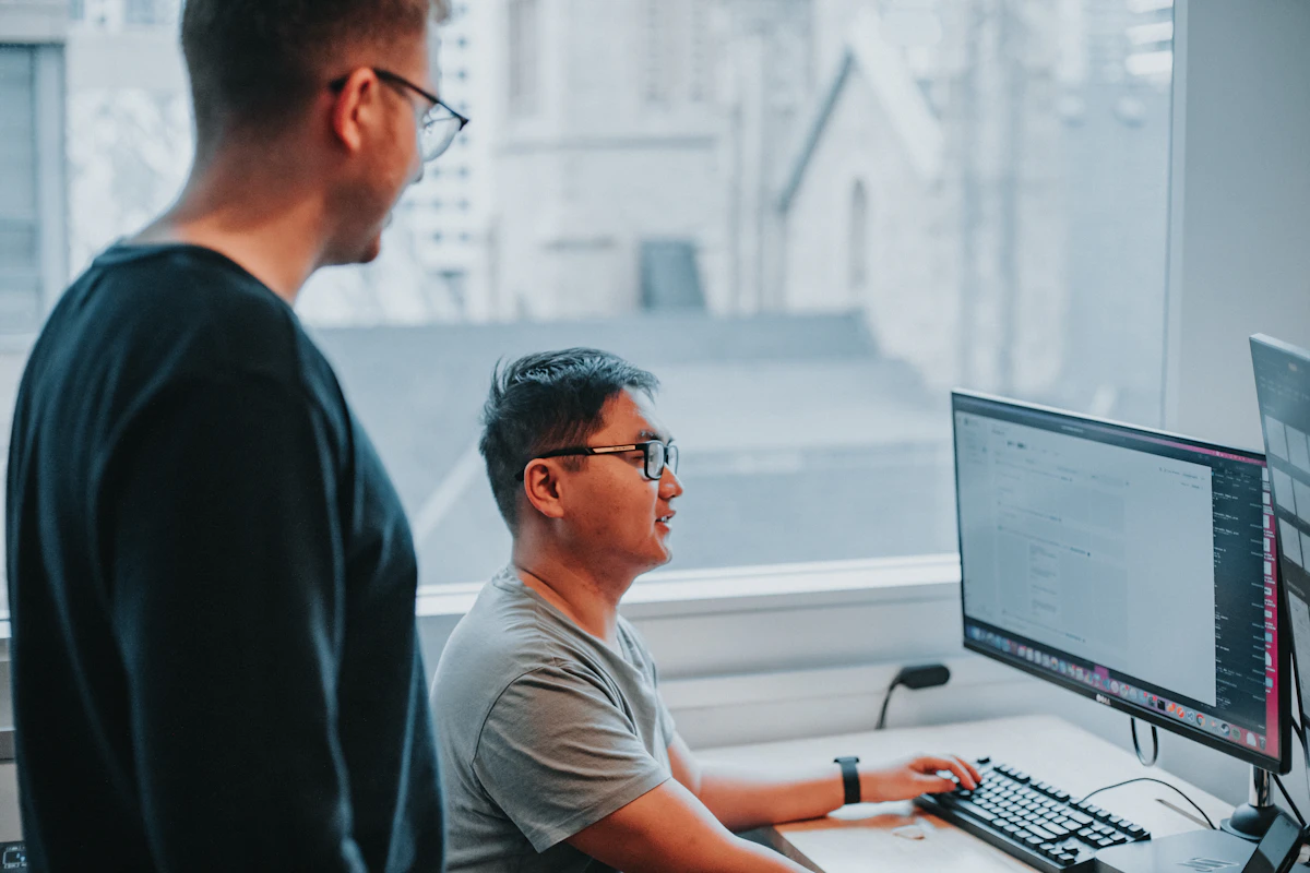 A man in glasses works at a desktop computer by a bright window while a colleague stands at his shoulder watching the screen
