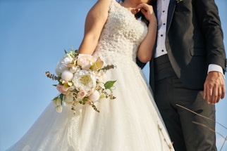 A bride and groom stand together, with the bride holding a bouquet of white and pastel-colored flowers. The bride wears a white, intricately embroidered wedding gown, while the groom is dressed in a black tuxedo with a white shirt and vest. The image is captured from a low angle, emphasizing the blue sky in the background.