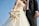 A bride and groom stand together, with the bride holding a bouquet of white and pastel-colored flowers. The bride wears a white, intricately embroidered wedding gown, while the groom is dressed in a black tuxedo with a white shirt and vest. The image is captured from a low angle, emphasizing the blue sky in the background.