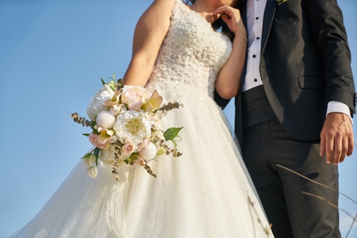 A bride and groom stand together, with the bride holding a bouquet of white and pastel-colored flowers. The bride wears a white, intricately embroidered wedding gown, while the groom is dressed in a black tuxedo with a white shirt and vest. The image is captured from a low angle, emphasizing the blue sky in the background.