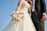 A bride and groom stand together, with the bride holding a bouquet of white and pastel-colored flowers. The bride wears a white, intricately embroidered wedding gown, while the groom is dressed in a black tuxedo with a white shirt and vest. The image is captured from a low angle, emphasizing the blue sky in the background.