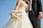 A bride and groom stand together, with the bride holding a bouquet of white and pastel-colored flowers. The bride wears a white, intricately embroidered wedding gown, while the groom is dressed in a black tuxedo with a white shirt and vest. The image is captured from a low angle, emphasizing the blue sky in the background.