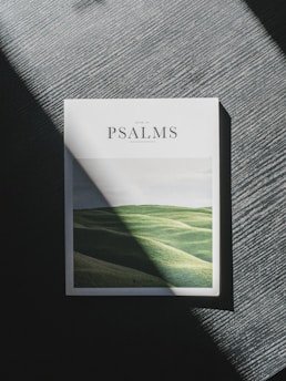 A book with a cover featuring the word 'Psalms' is placed on a textured surface. Sunlight casts a diagonal shadow, highlighting the image of rolling green hills on the book cover.