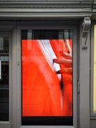 A digital display embedded in a framed structure shows a close-up image of a hand gently touching or inspecting an object with a textured surface. The primary background color is a bold red, and the scene is partially reflected on the glass, showcasing subtle elements of the surrounding exterior environment.
