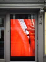 A digital display embedded in a framed structure shows a close-up image of a hand gently touching or inspecting an object with a textured surface. The primary background color is a bold red, and the scene is partially reflected on the glass, showcasing subtle elements of the surrounding exterior environment.