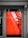 A digital display embedded in a framed structure shows a close-up image of a hand gently touching or inspecting an object with a textured surface. The primary background color is a bold red, and the scene is partially reflected on the glass, showcasing subtle elements of the surrounding exterior environment.