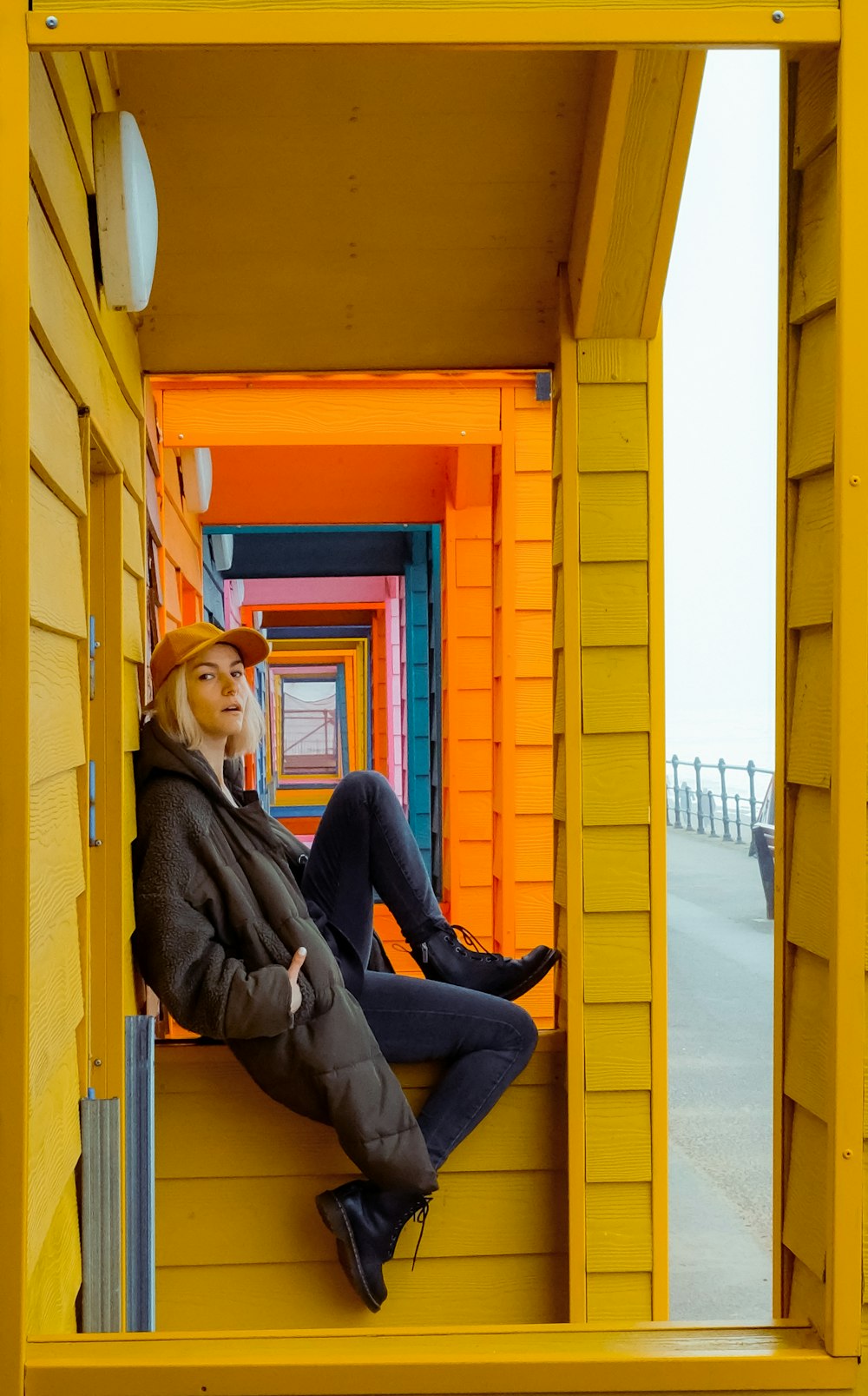 woman in brown jacket and blue denim jeans sitting on yellow wooden door during daytime